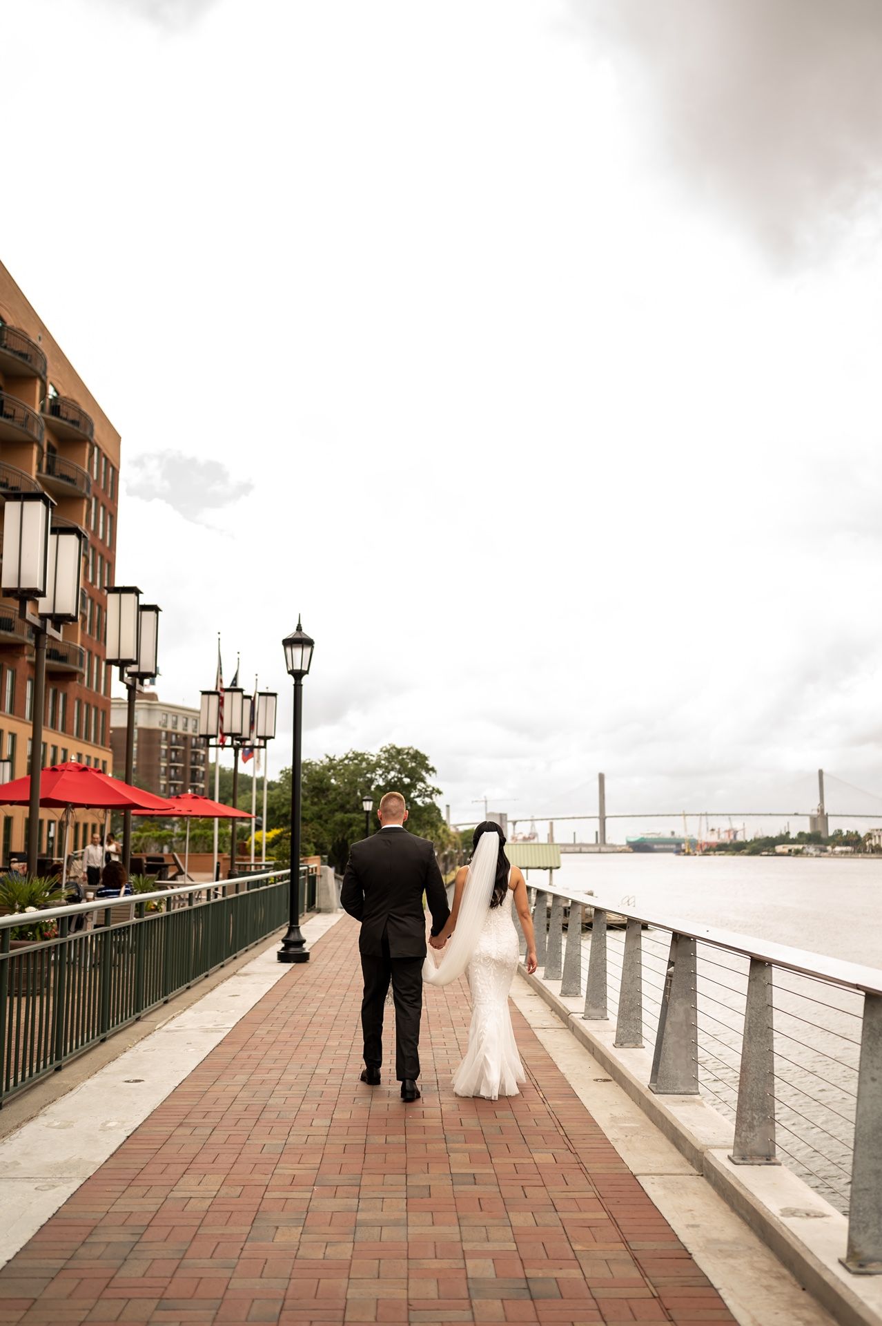Two people walk along a brick waterfront promenade with classic lamp posts and a bridge visible in the distance.