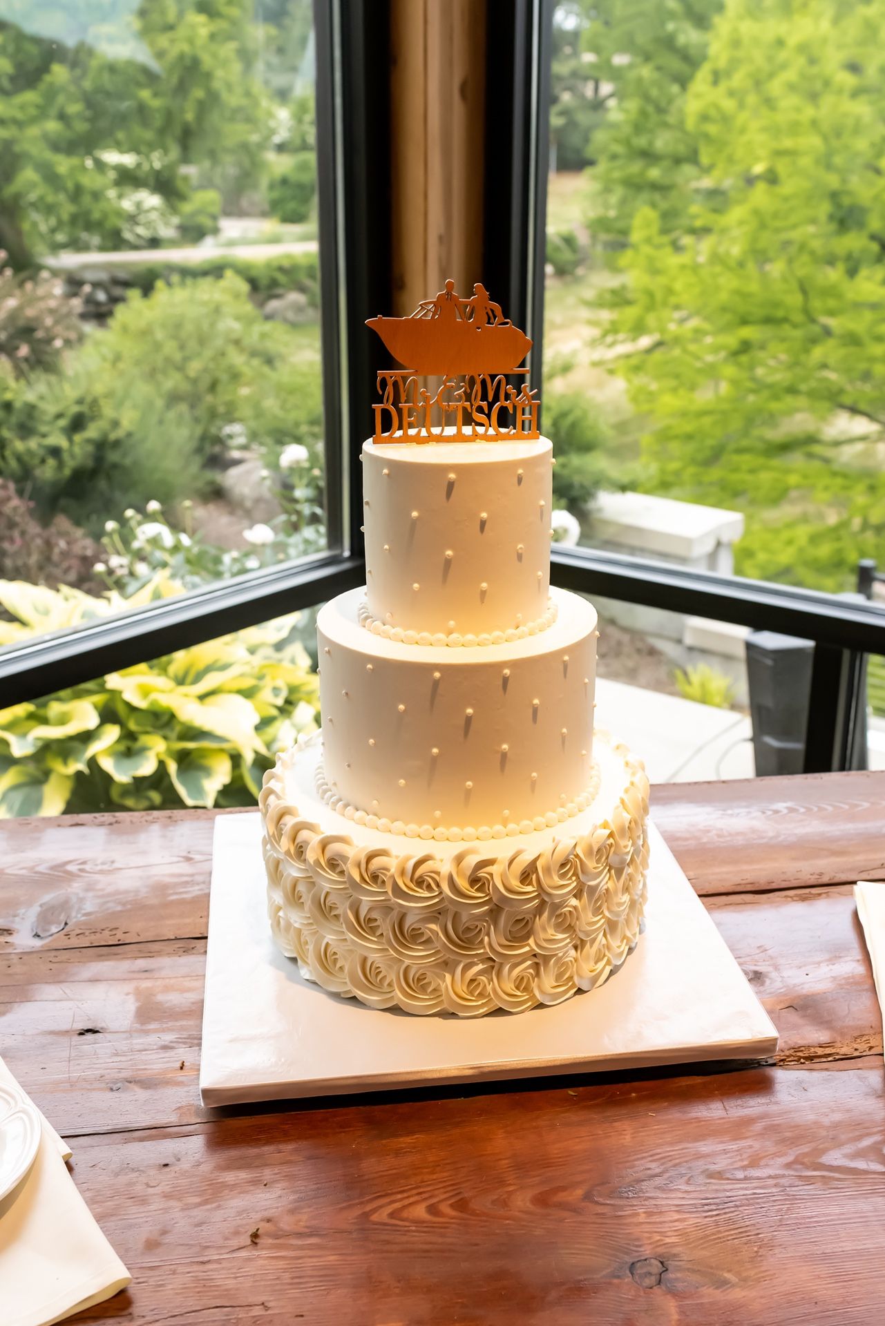 Elegant three-tiered white wedding cake with polka dots and gold rosette details displayed by a window with garden view.