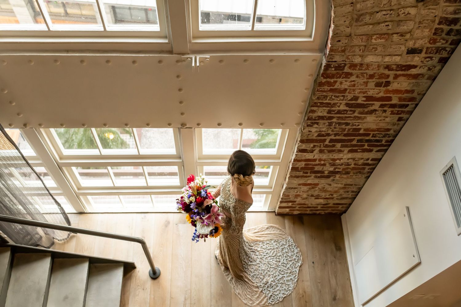 A person in formal attire stands alone on a spiral staircase landing holding a vibrant floral bouquet near tall windows.