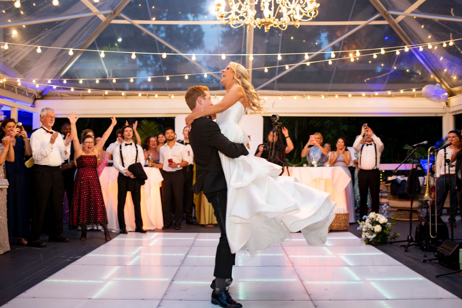 Bride and groom share first dance under elegant tent with string lights, guests cheering in background at wedding reception.