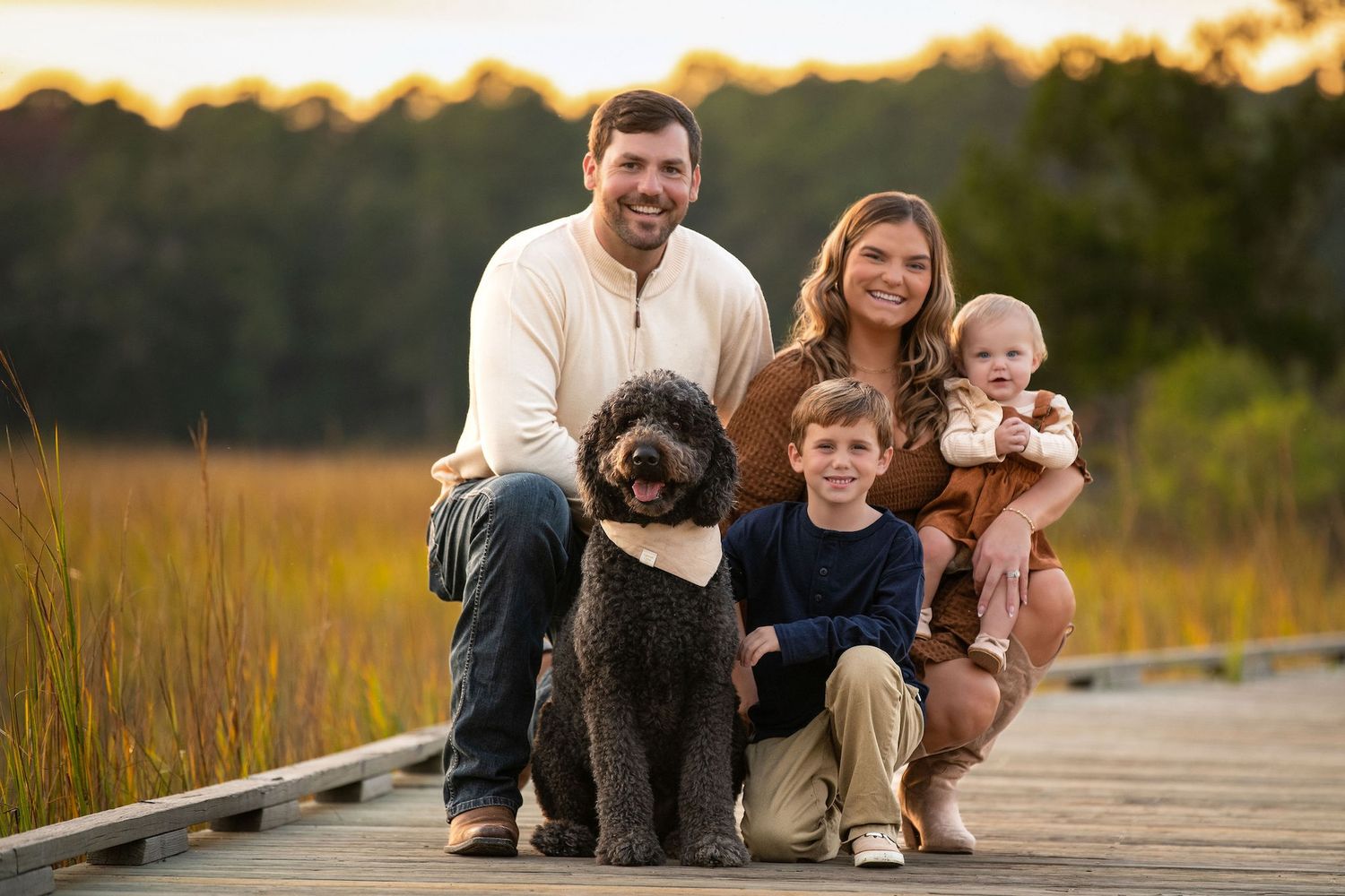 Family of four with a dog posing on a wooden boardwalk surrounded by golden marsh grass at sunset.