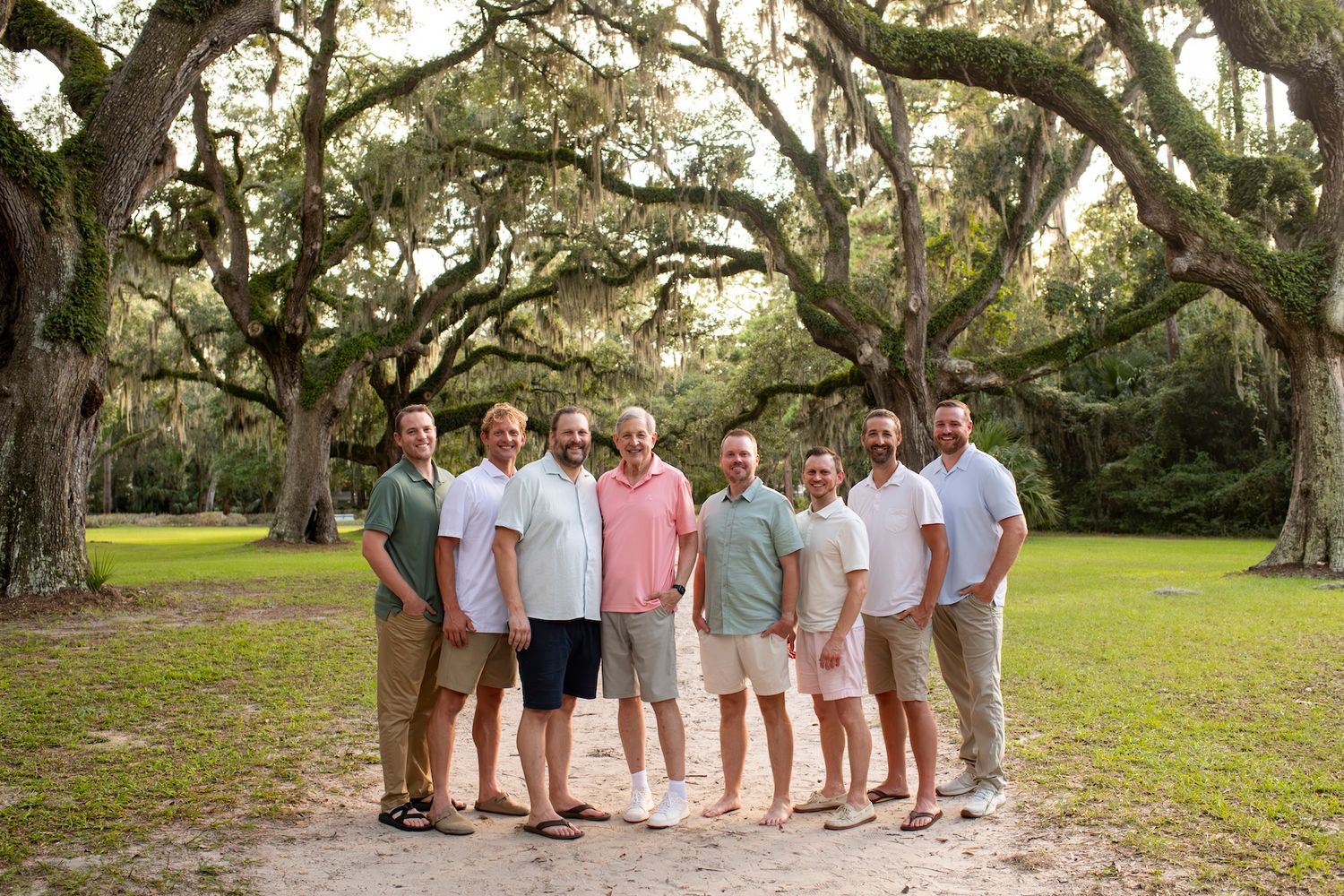 Group of seven men smiling together on a sandy path under sprawling moss-covered oak trees outdoors.