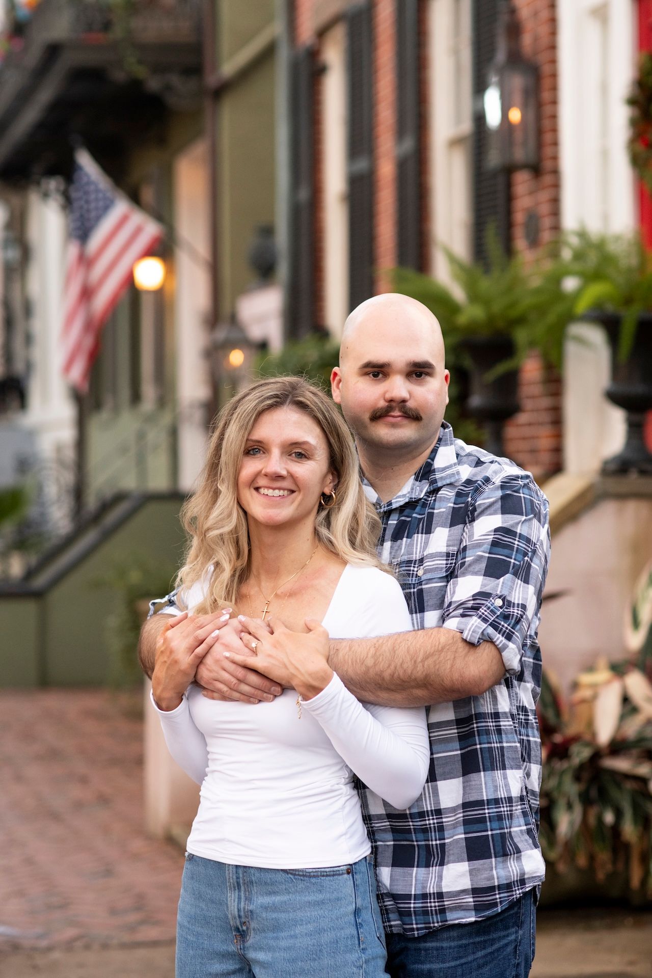 A couple in casual attire poses together on a brick sidewalk in front of historic buildings.