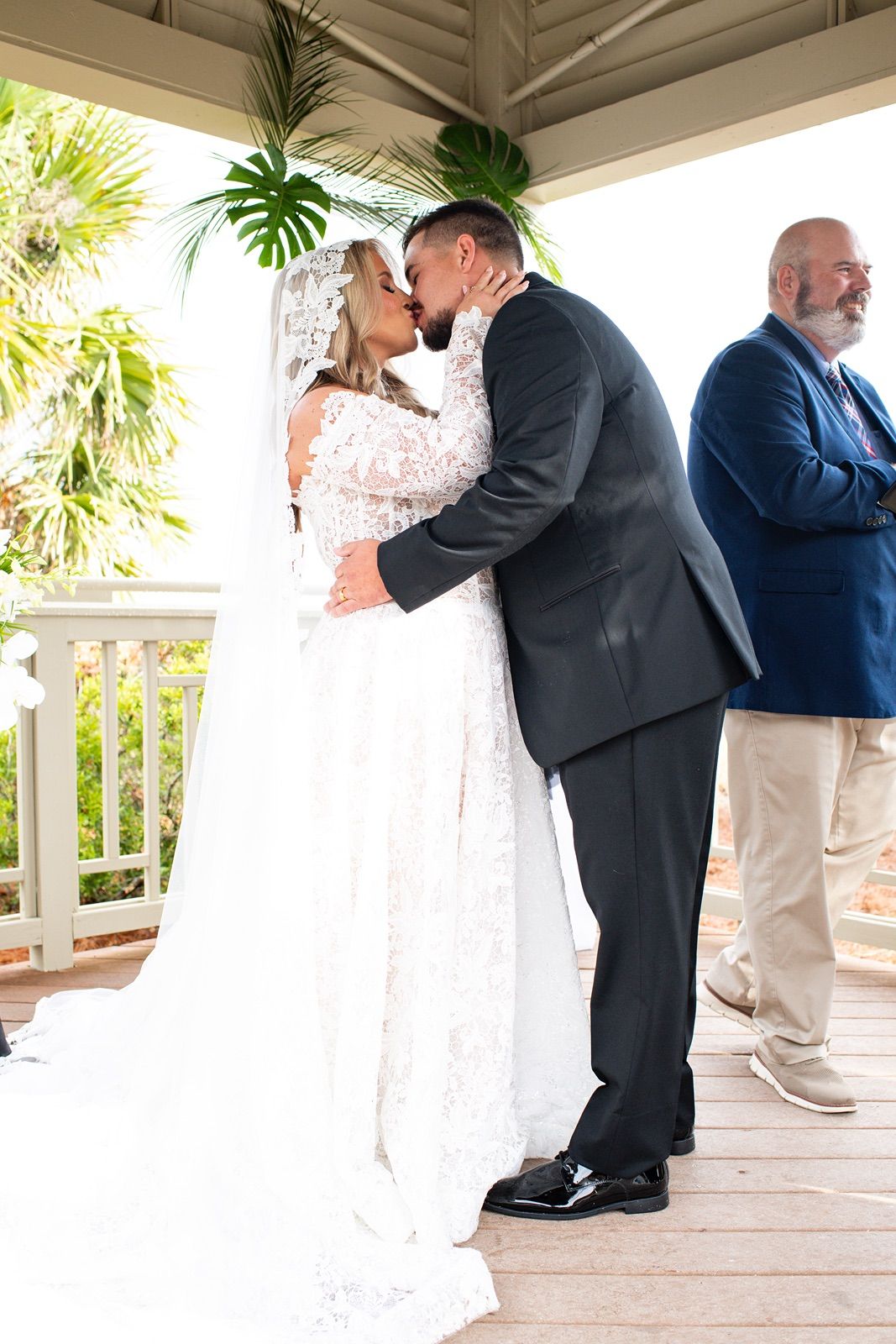 Wedding couple shares a romantic kiss on a covered white porch with tropical plants in the background.