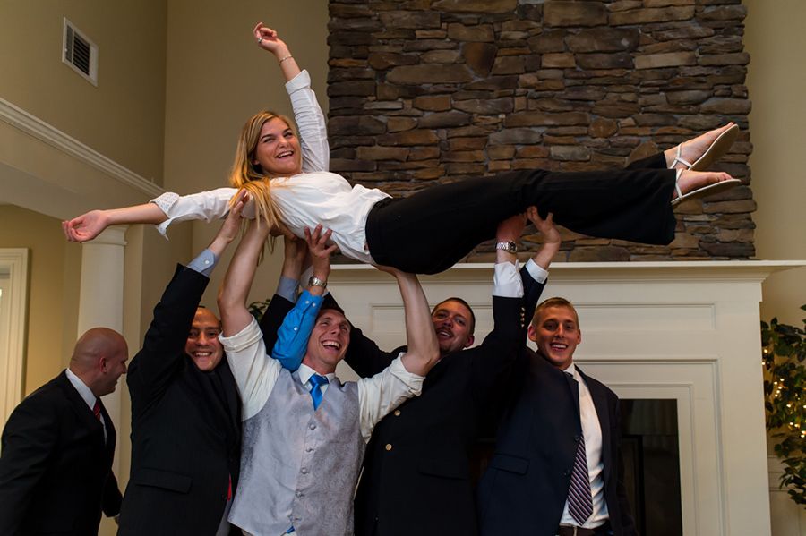 A celebratory scene shows wedding party members lifting and holding someone horizontally in front of a stone fireplace.