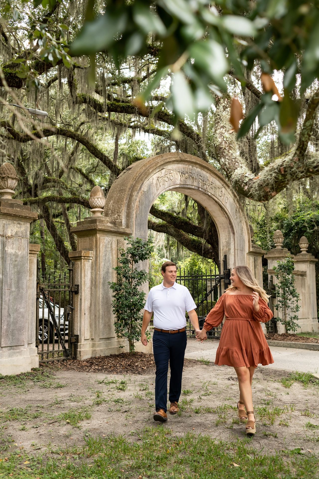 A couple walking together beneath a beautiful stone archway in a lush garden with Spanish moss hanging overhead.