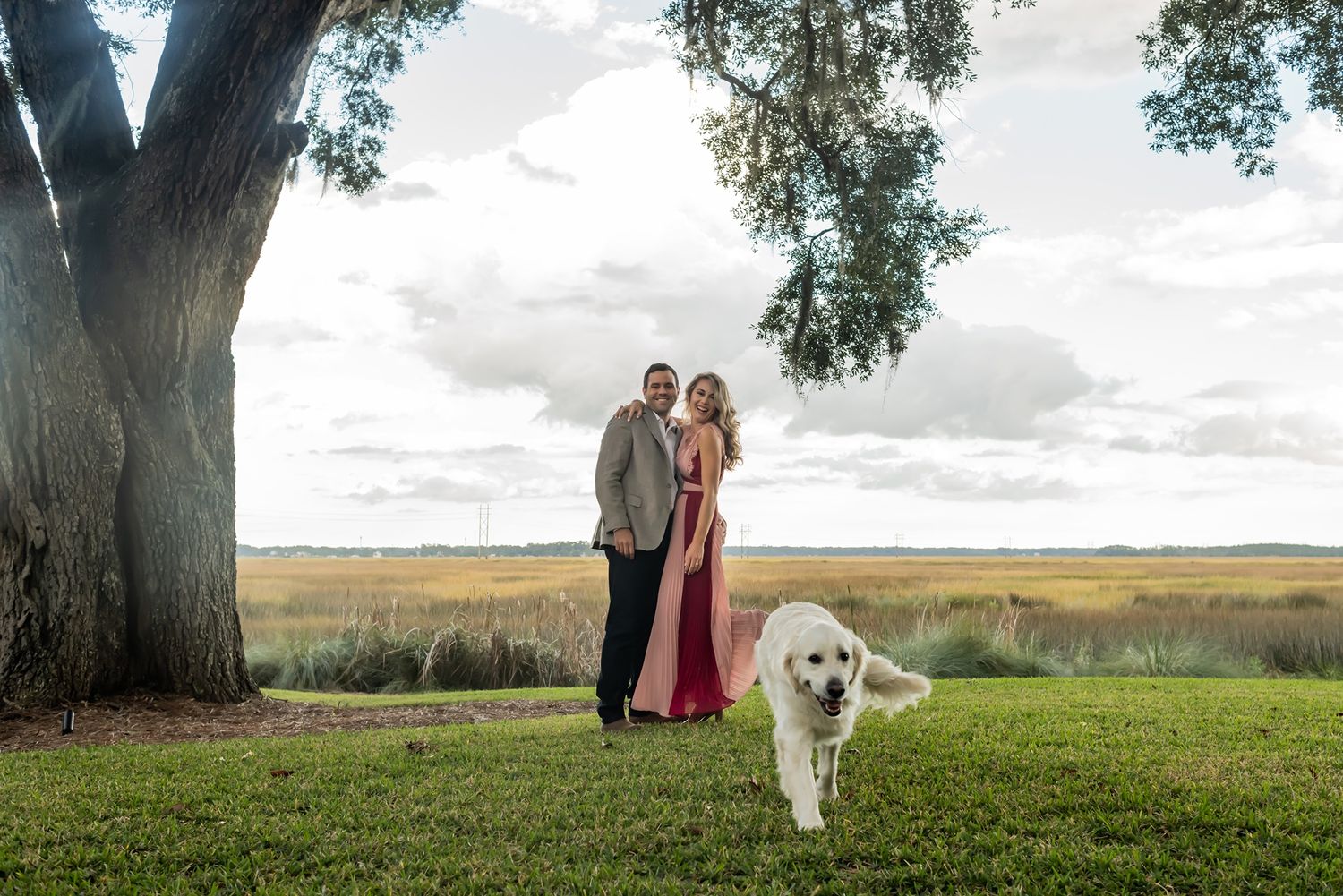 A couple and their golden retriever pose together under a large tree in a scenic countryside setting during sunset.
