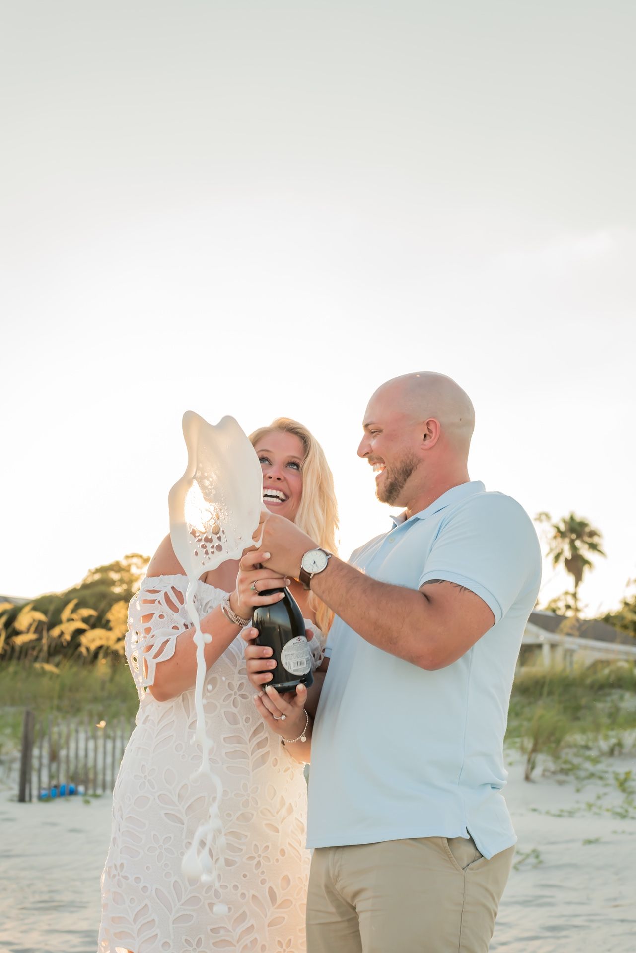 Couple celebrating with champagne and white sunhat on beach at sunset with palm trees in background.