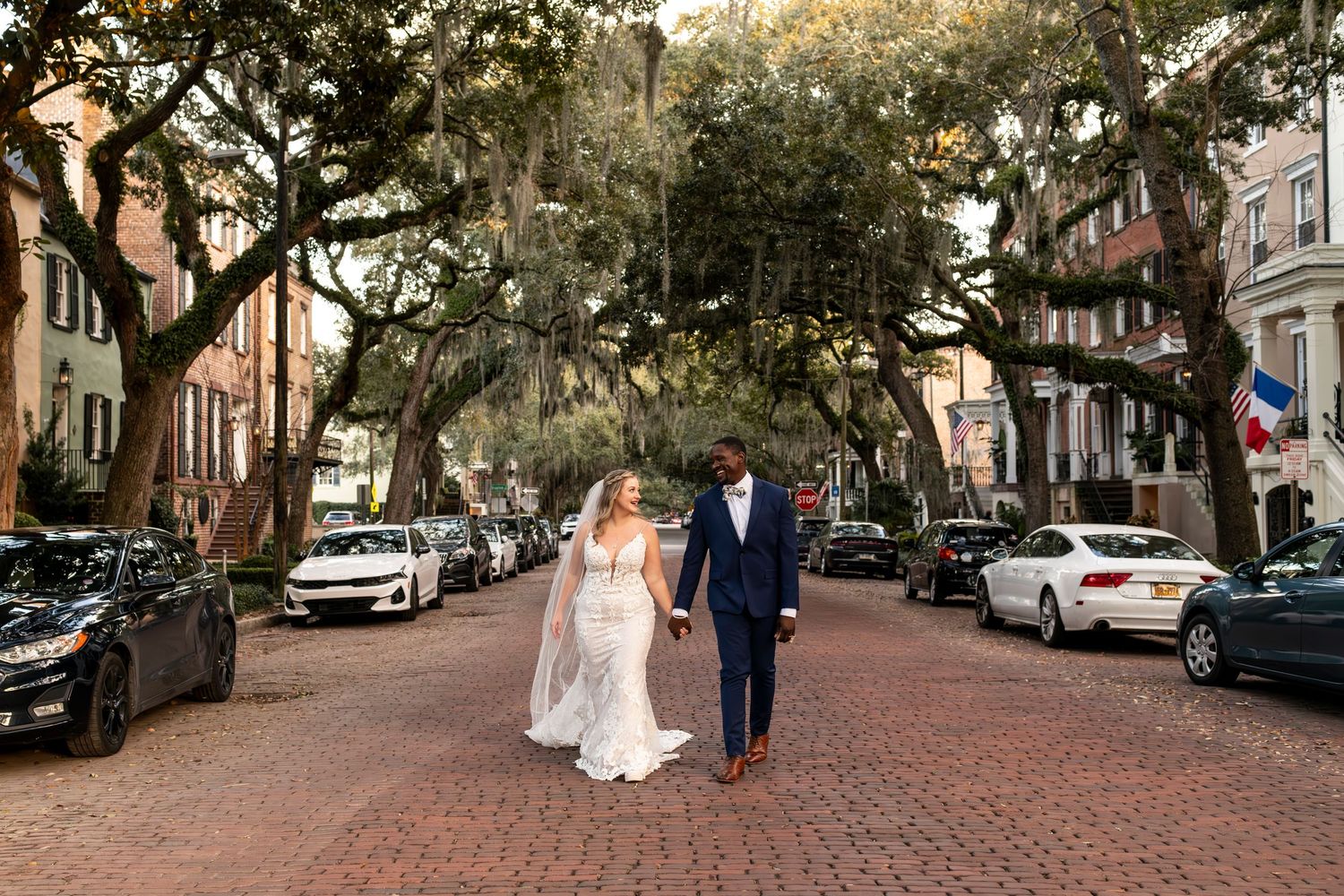 A couple walks down a historic brick street lined with Spanish moss-draped oak trees and parked cars in Savannah Georgia.