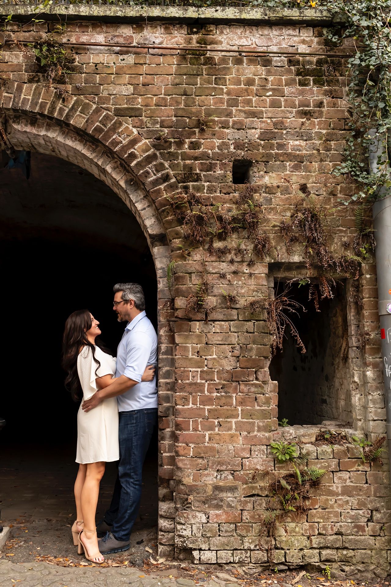 Two people embrace in front of a rustic brick archway covered in vines at a historic building location.