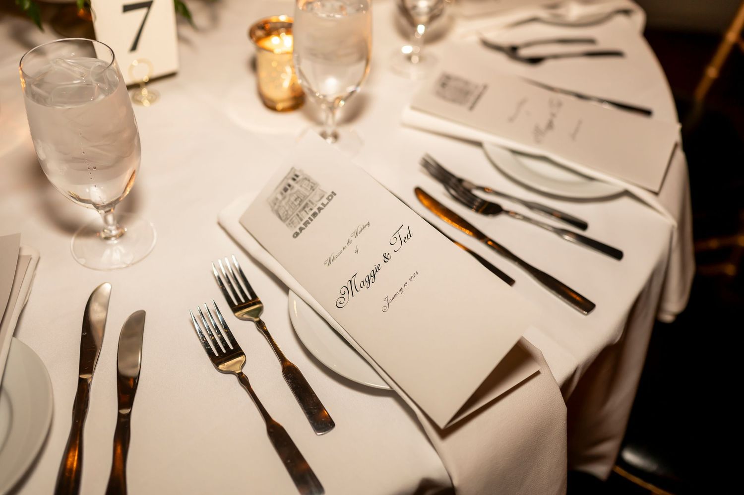 A formal dinner table setting with white linens, silverware, glassware and menu cards arranged at each place.