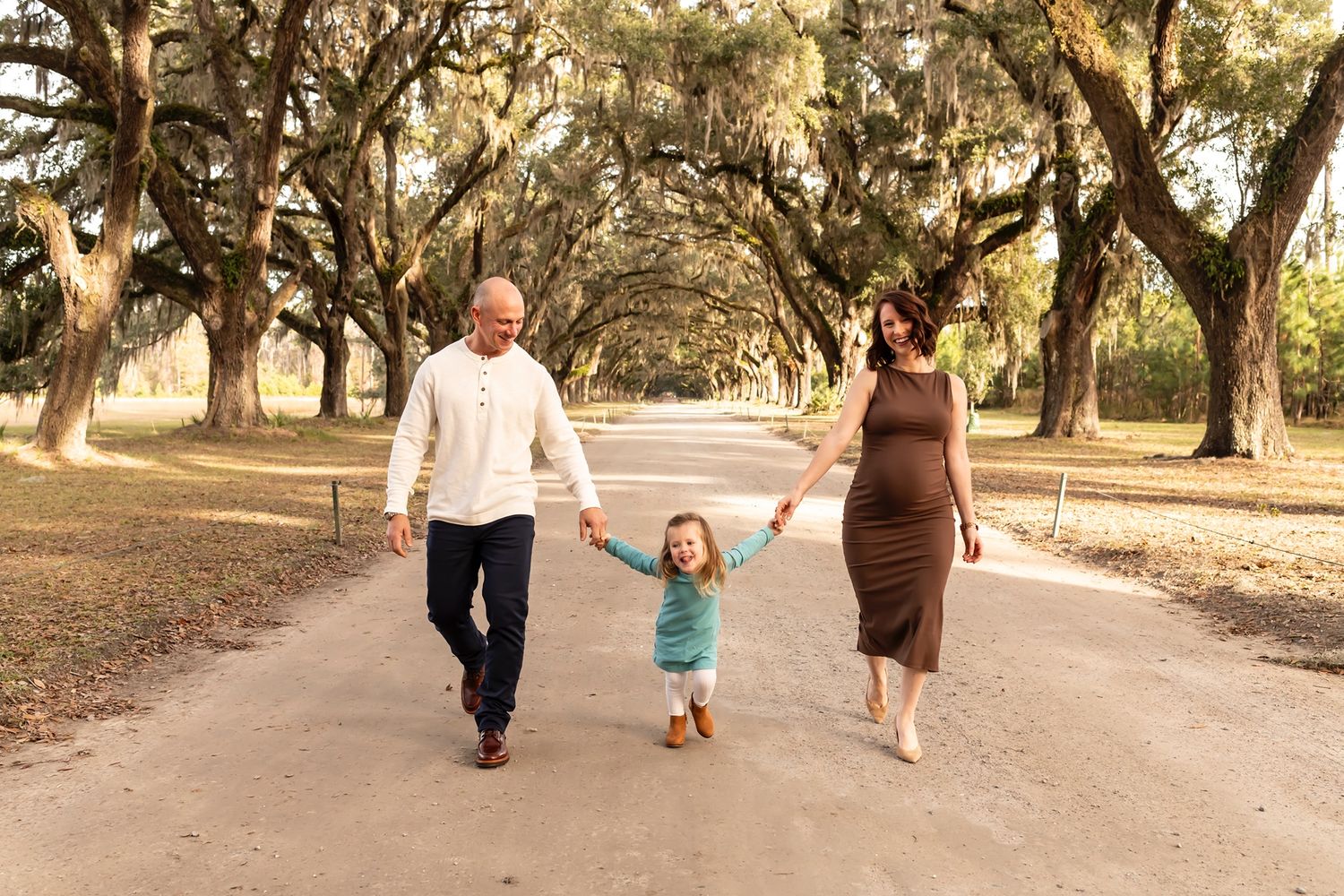 A child walks while holding hands with two adults down a path lined with majestic oak trees draped in Spanish moss.