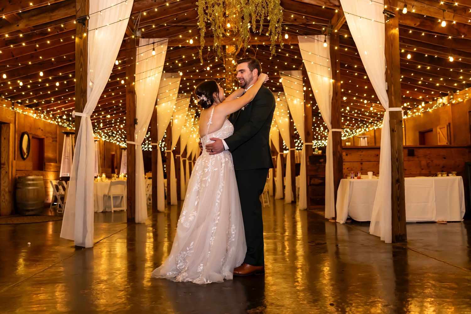 A romantic dance takes place under twinkling lights in a rustic barn venue during an elegant wedding reception.