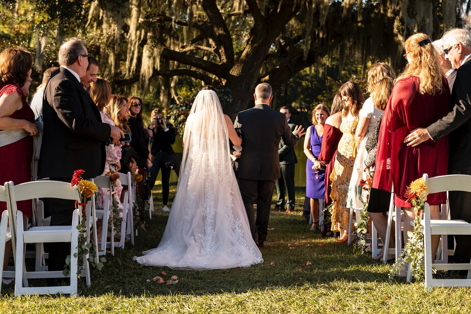 Outdoor wedding ceremony under moss-draped trees with guests seated on white chairs along a laneway decorated with flowers.