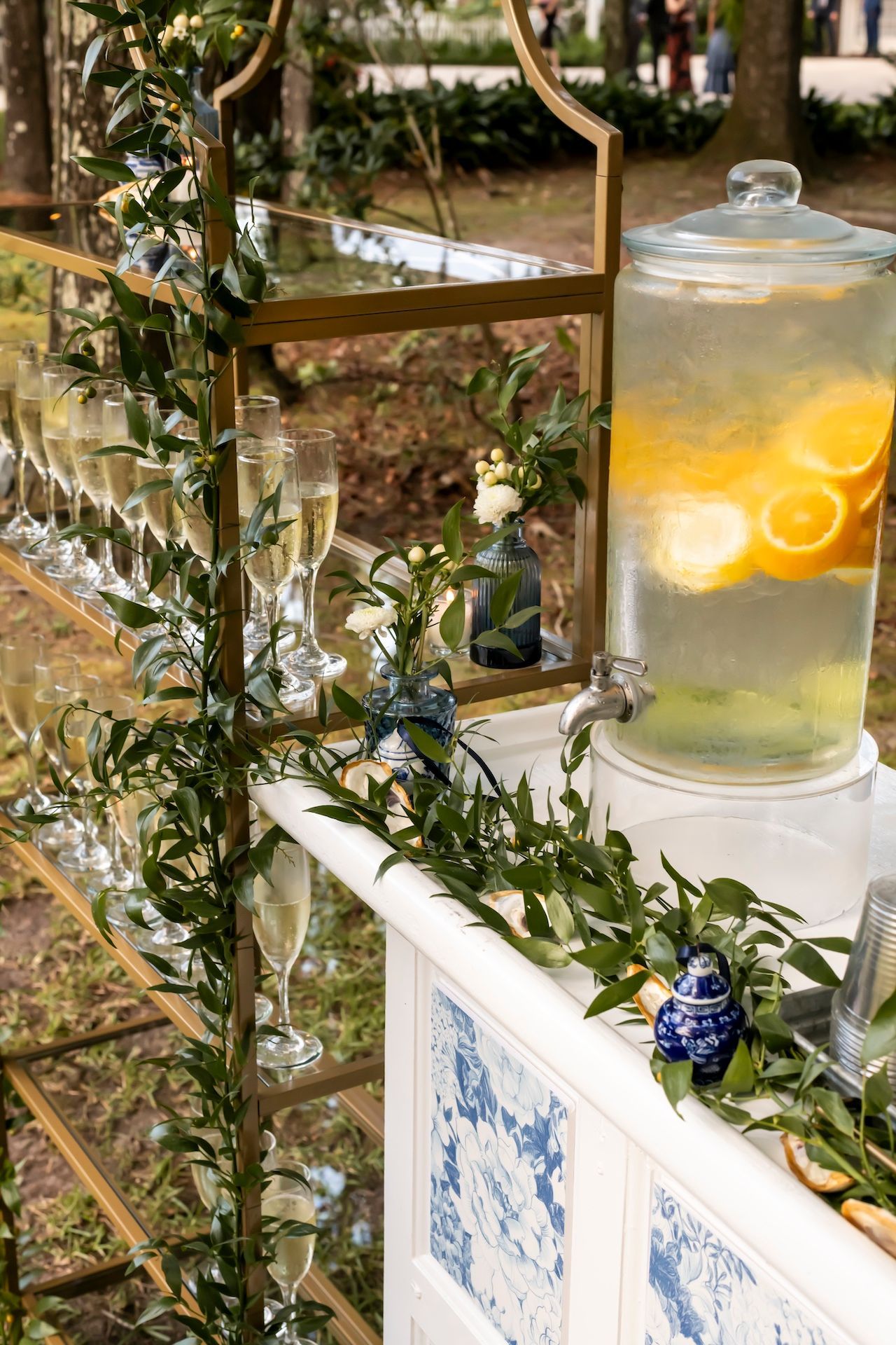 Glass beverage dispenser with citrus slices sits on a decorative white bar adorned with greenery and blue accents at an outdoor event.