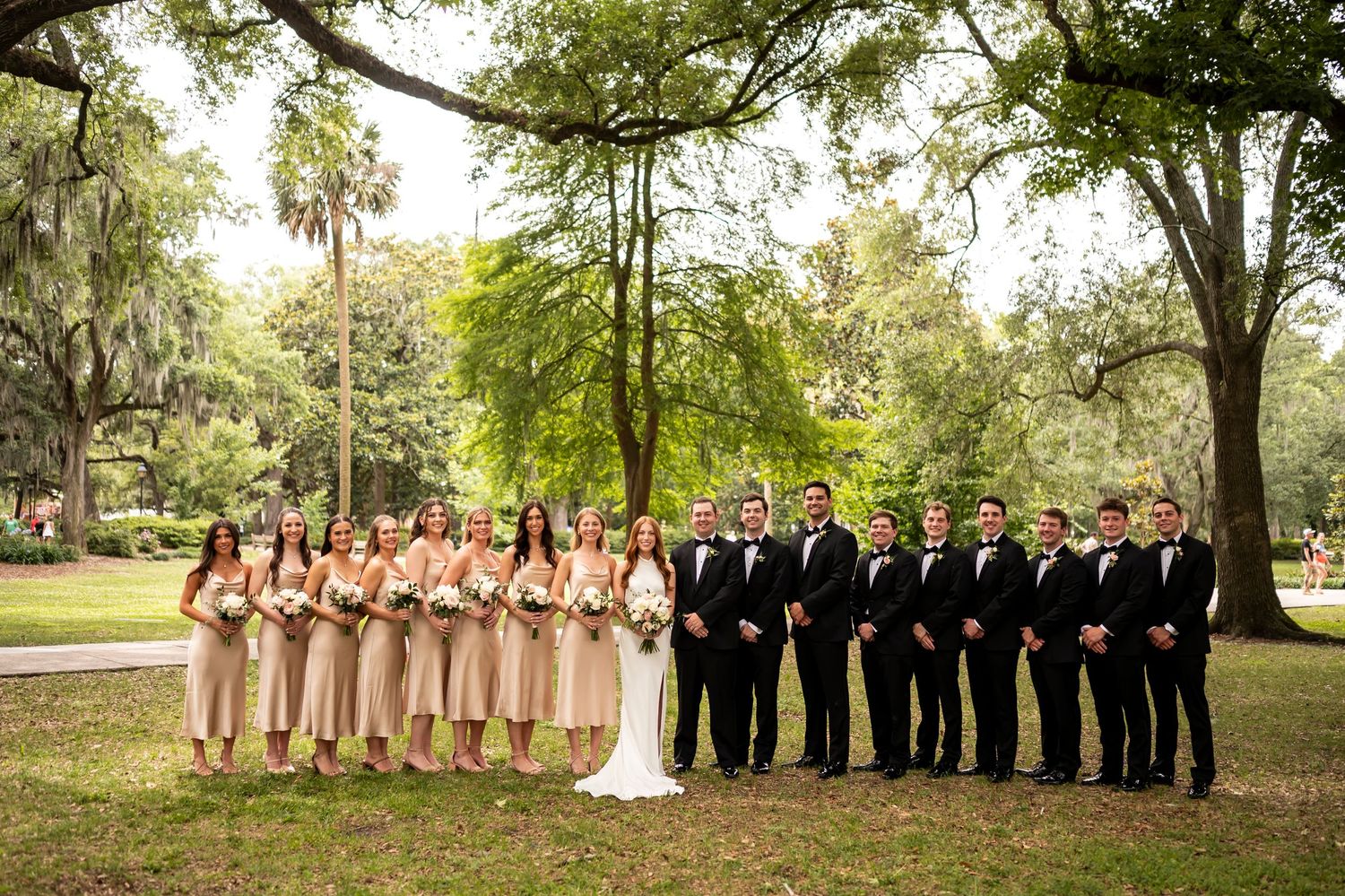 A wedding party poses outdoors under large oak trees draped with Spanish moss in a garden setting.
