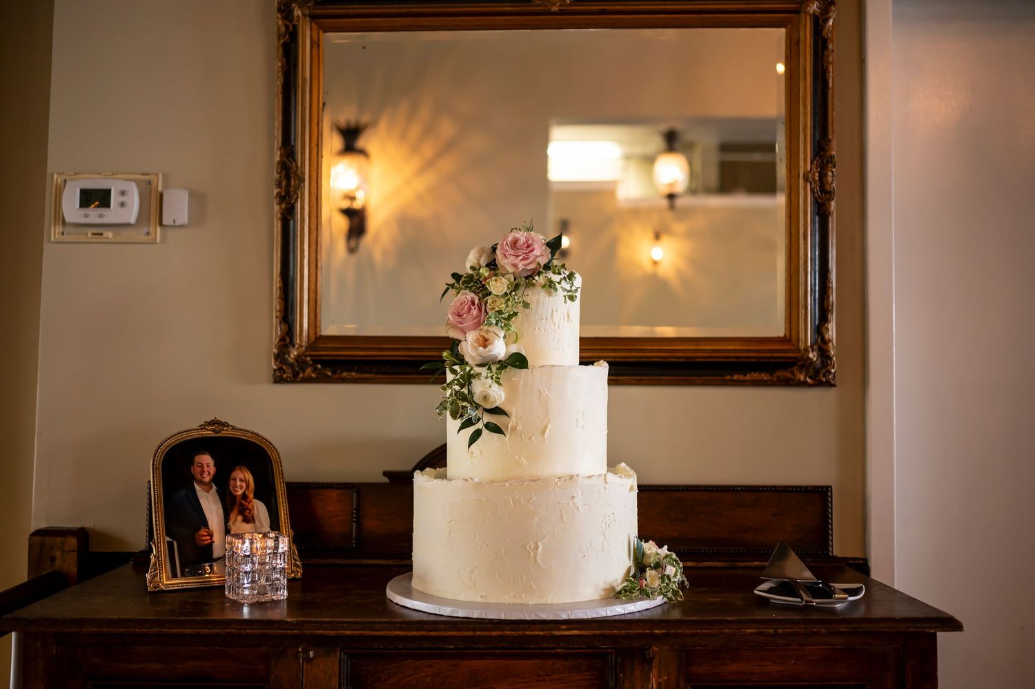Elegant three-tiered white wedding cake with floral decorations displayed on dark wooden table beneath ornate gold mirror.