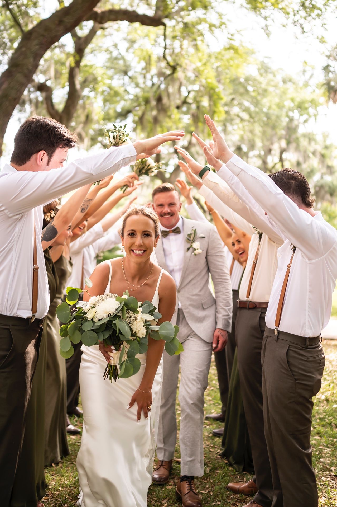 Wedding couple and bridal party celebrate under a tree arch as guests form a tunnel with raised arms for them to walk through.