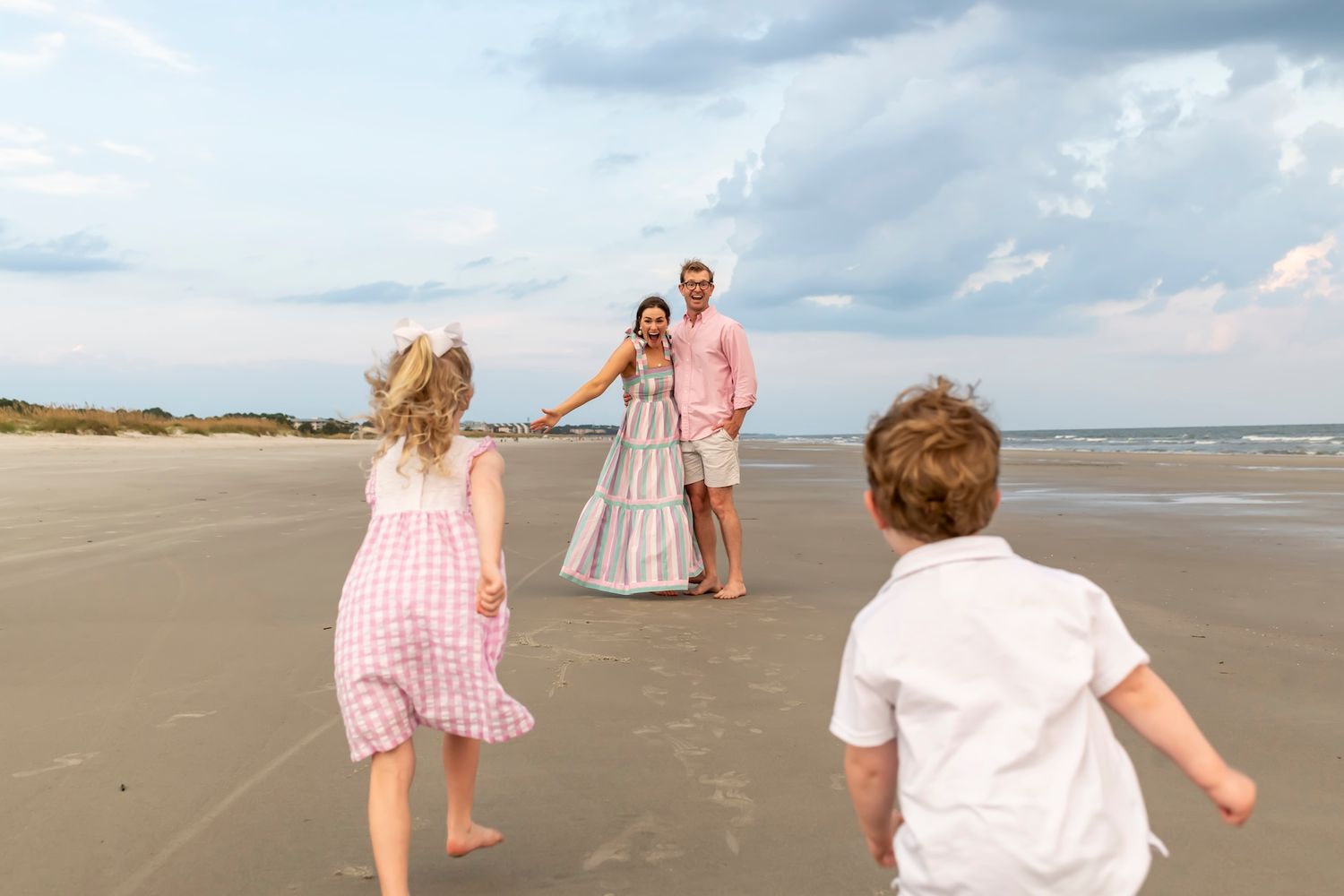 Children in pink and white outfits run toward family members on a serene beach during a cloudy summer day.