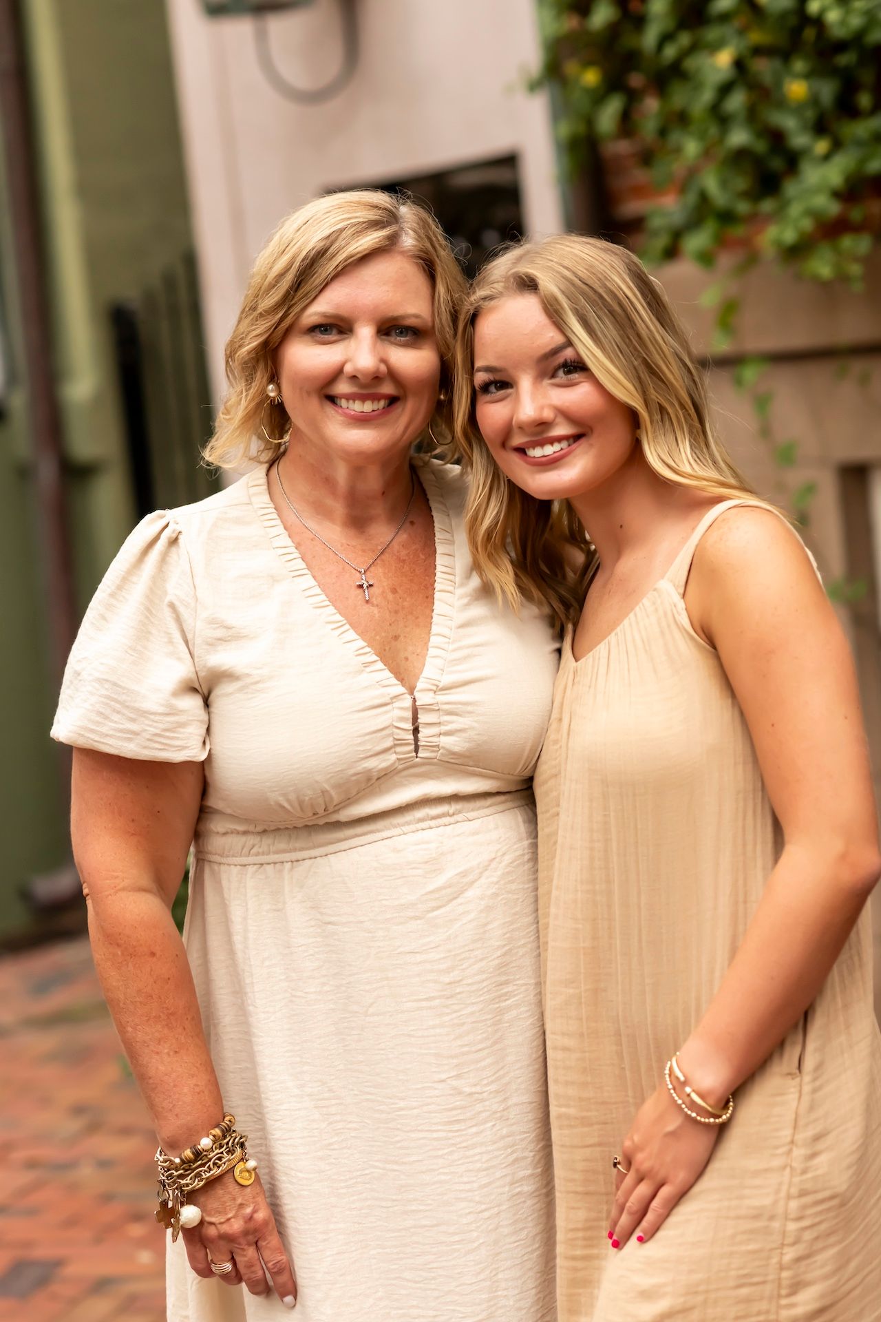 Two people wearing cream-colored dresses pose together on a brick patio with greenery in the background.