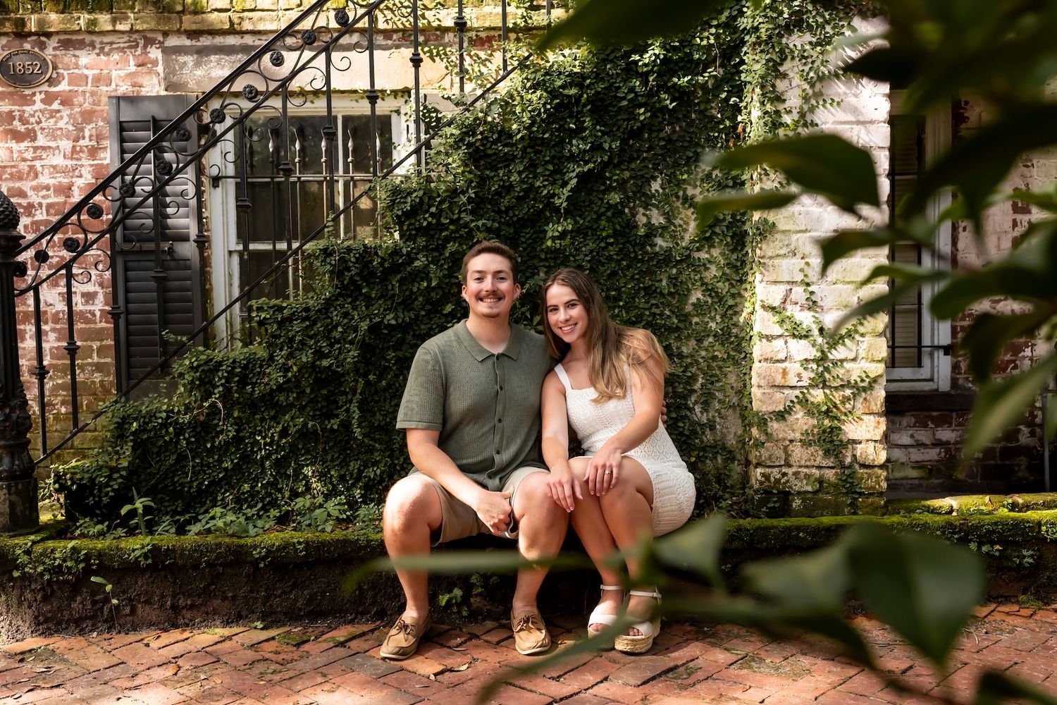 A couple smiles while sitting together on a brick stoop with ivy covered walls in the background.