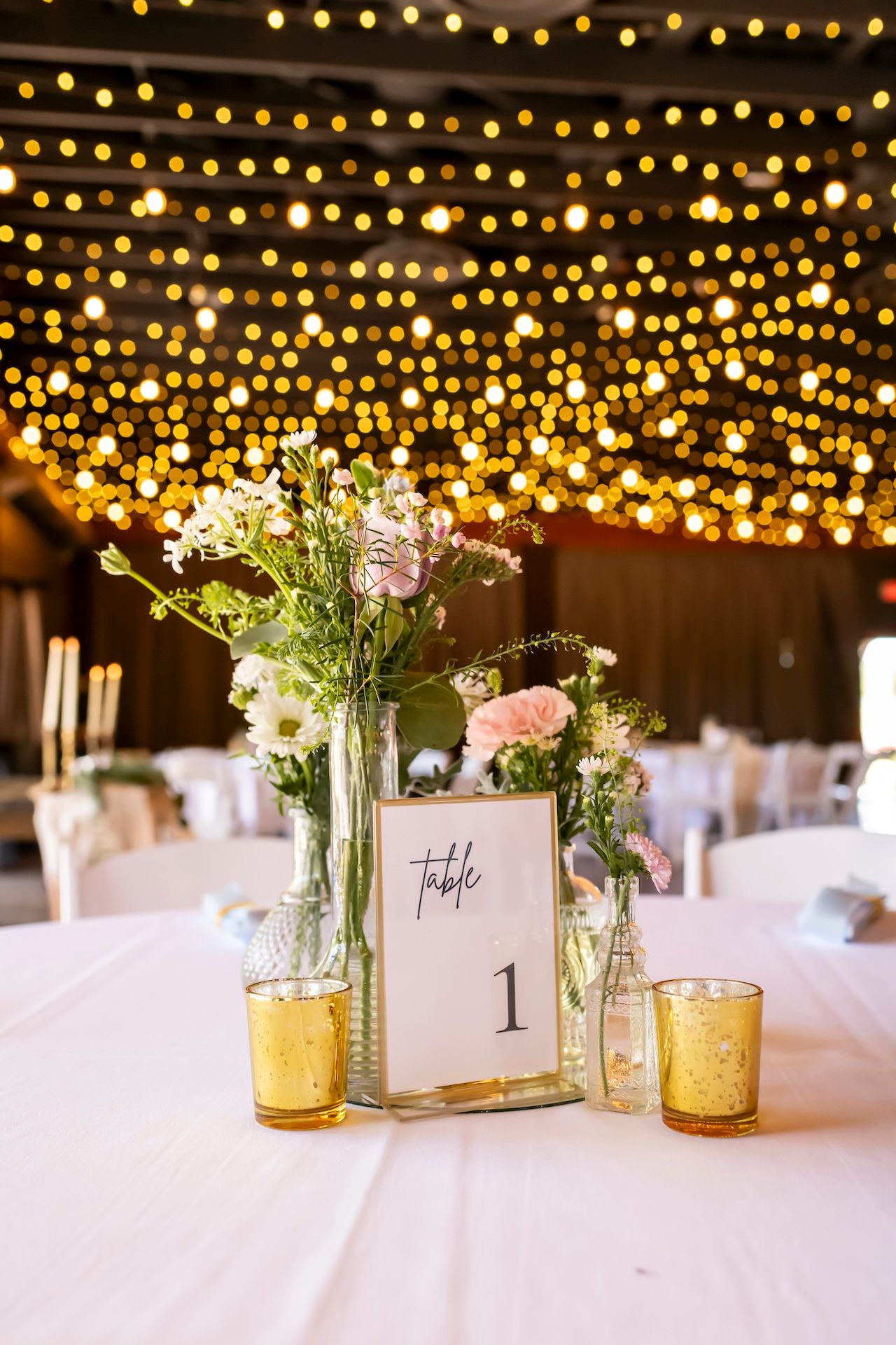 Wedding reception table decor with elegant floral centerpieces, gold candleholders and string lights overhead creating ambiance.
