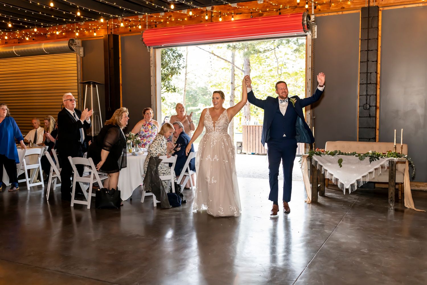 Wedding party dances under twinkling string lights in a rustic barn reception venue.