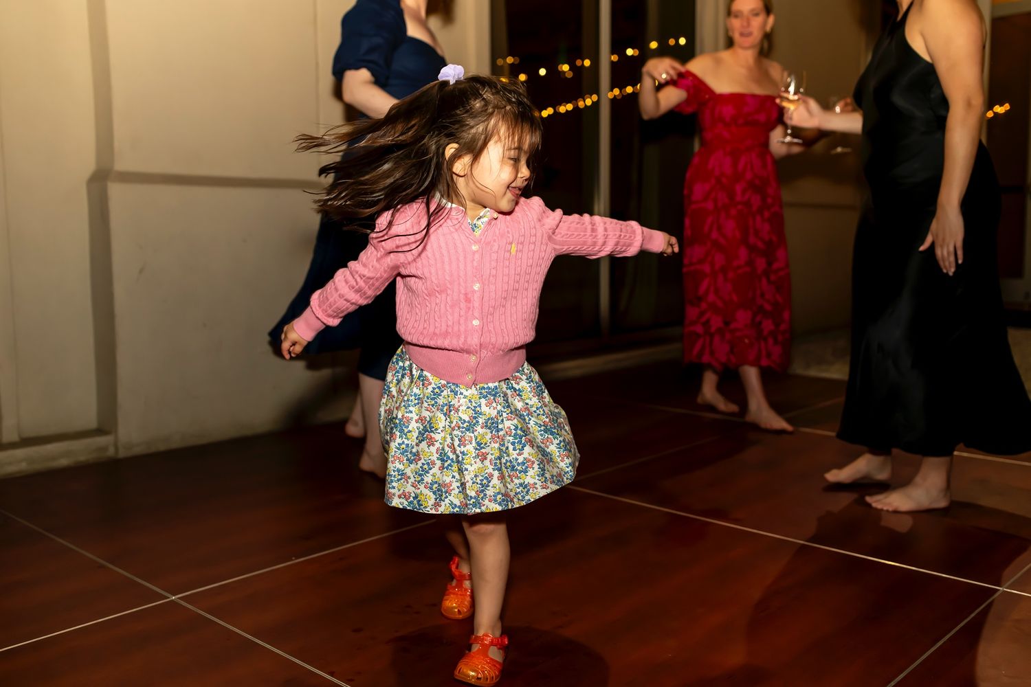 Young child spins joyfully on dance floor in pink shirt and patterned skirt during evening celebration.