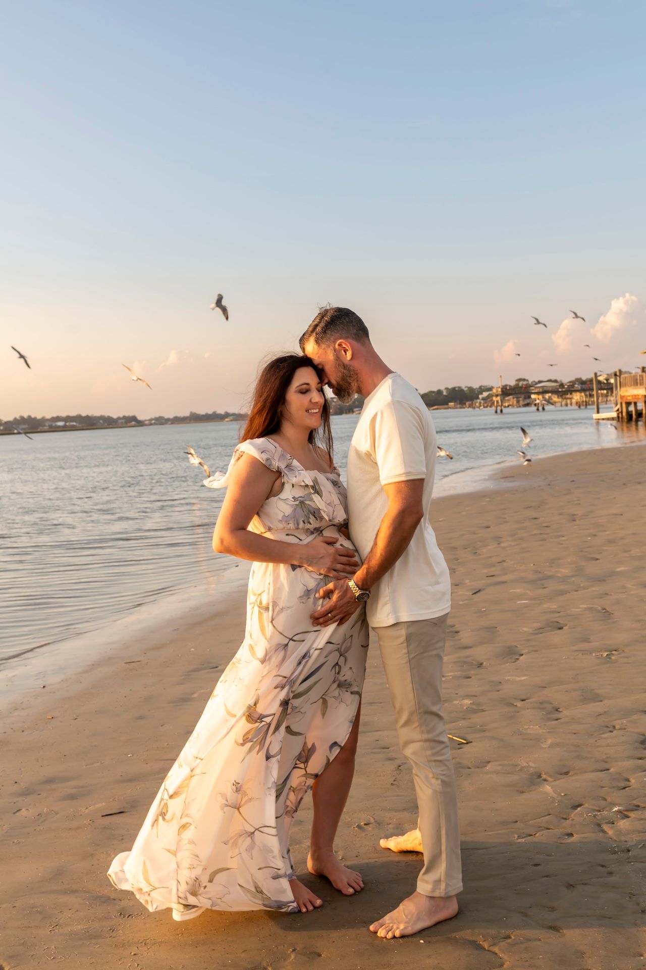 A romantic couple poses for a series of affectionate photos on a sandy beach at sunset with seagulls flying overhead.