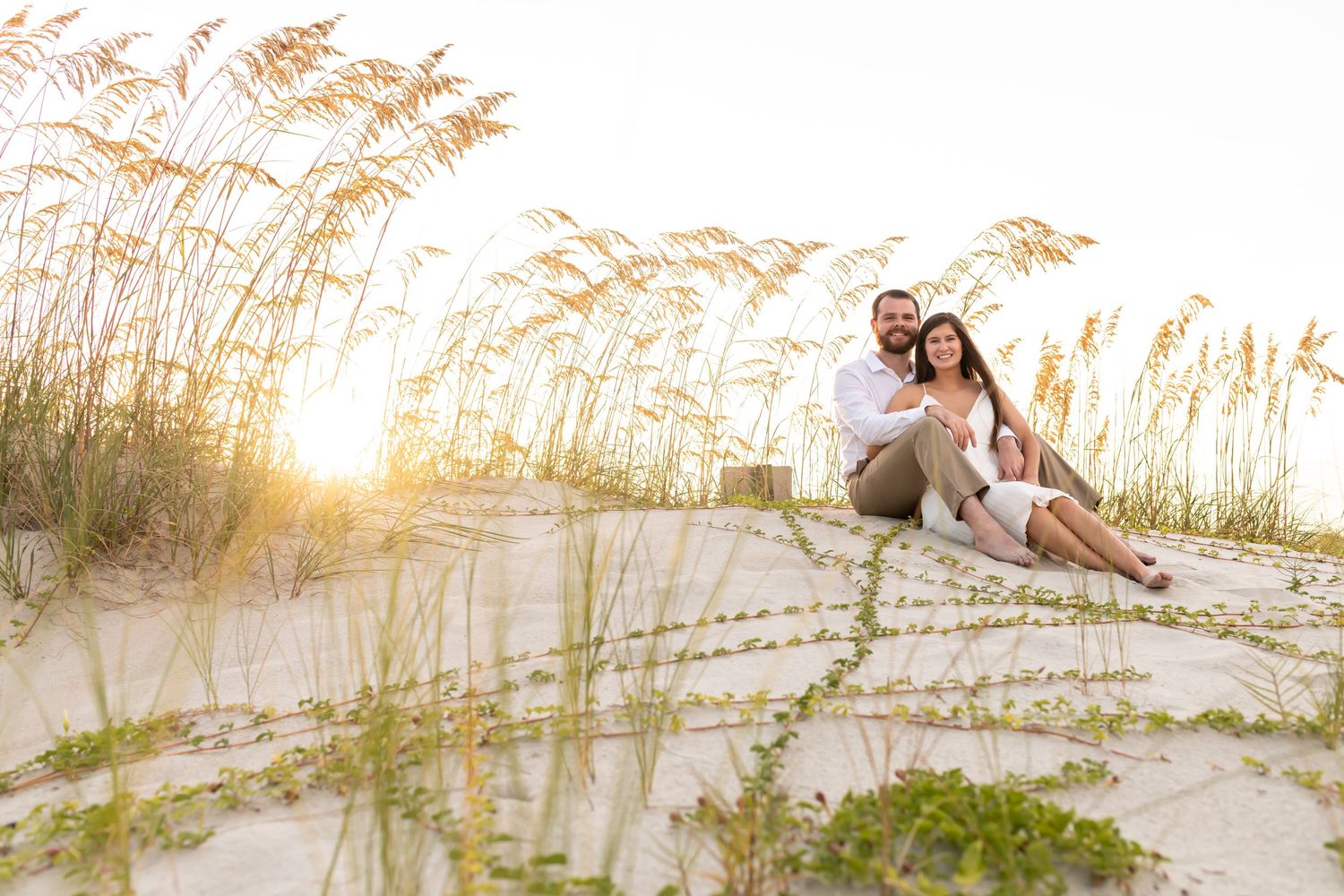 Romantic couple lying on sandy beach dunes with sea oats at sunset, creating silhouettes against golden sky.