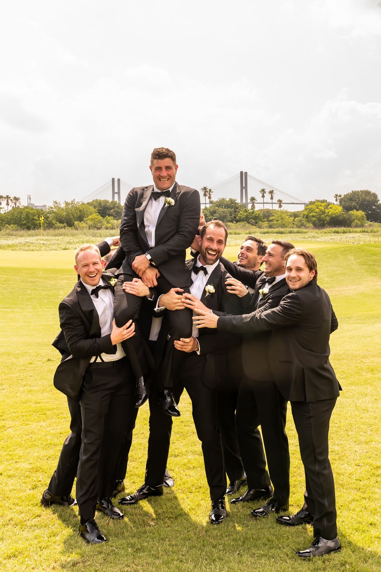 Groomsmen in black tuxedos lift and playfully carry someone on a green lawn with industrial buildings in background.