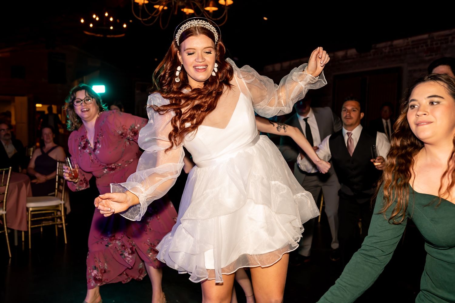 Person in a white party dress dances enthusiastically with other guests on a dimly lit dance floor at an event.