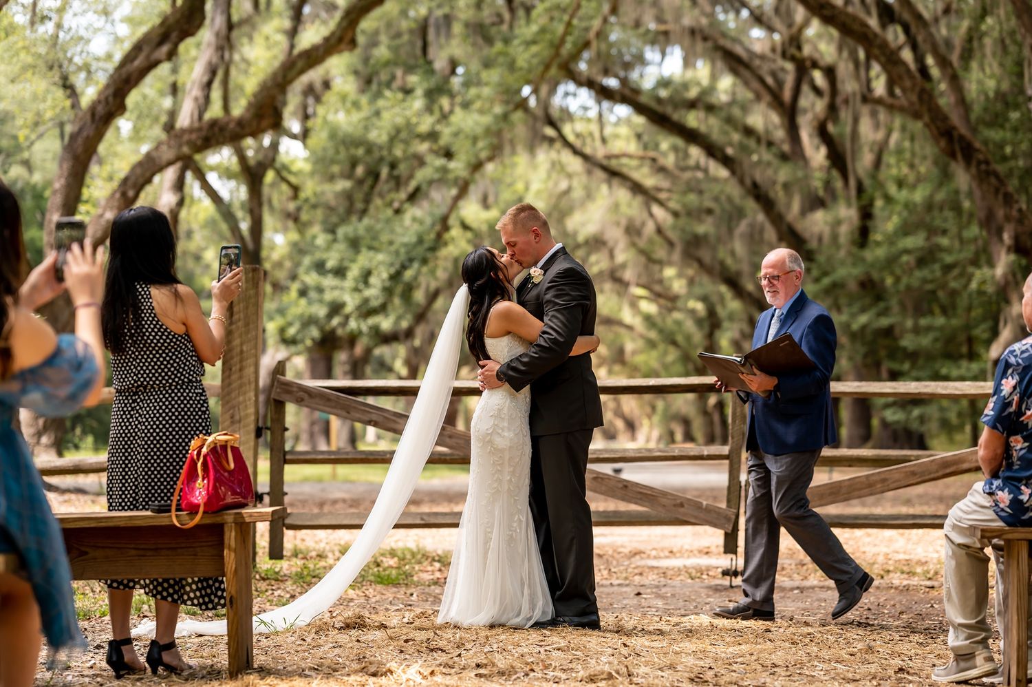An outdoor wedding ceremony under a canopy of oak trees as couple embraces with guests looking on.