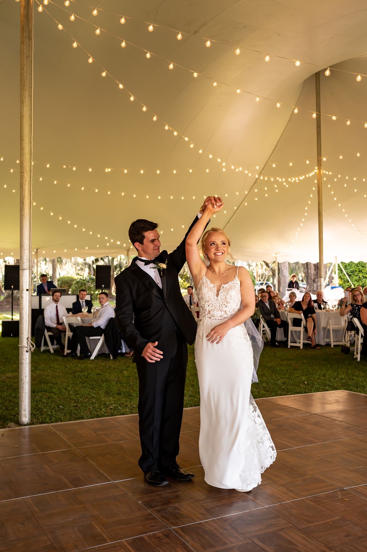 A couple shares their first dance on a wooden floor under a tent with twinkling string lights during an outdoor wedding reception.