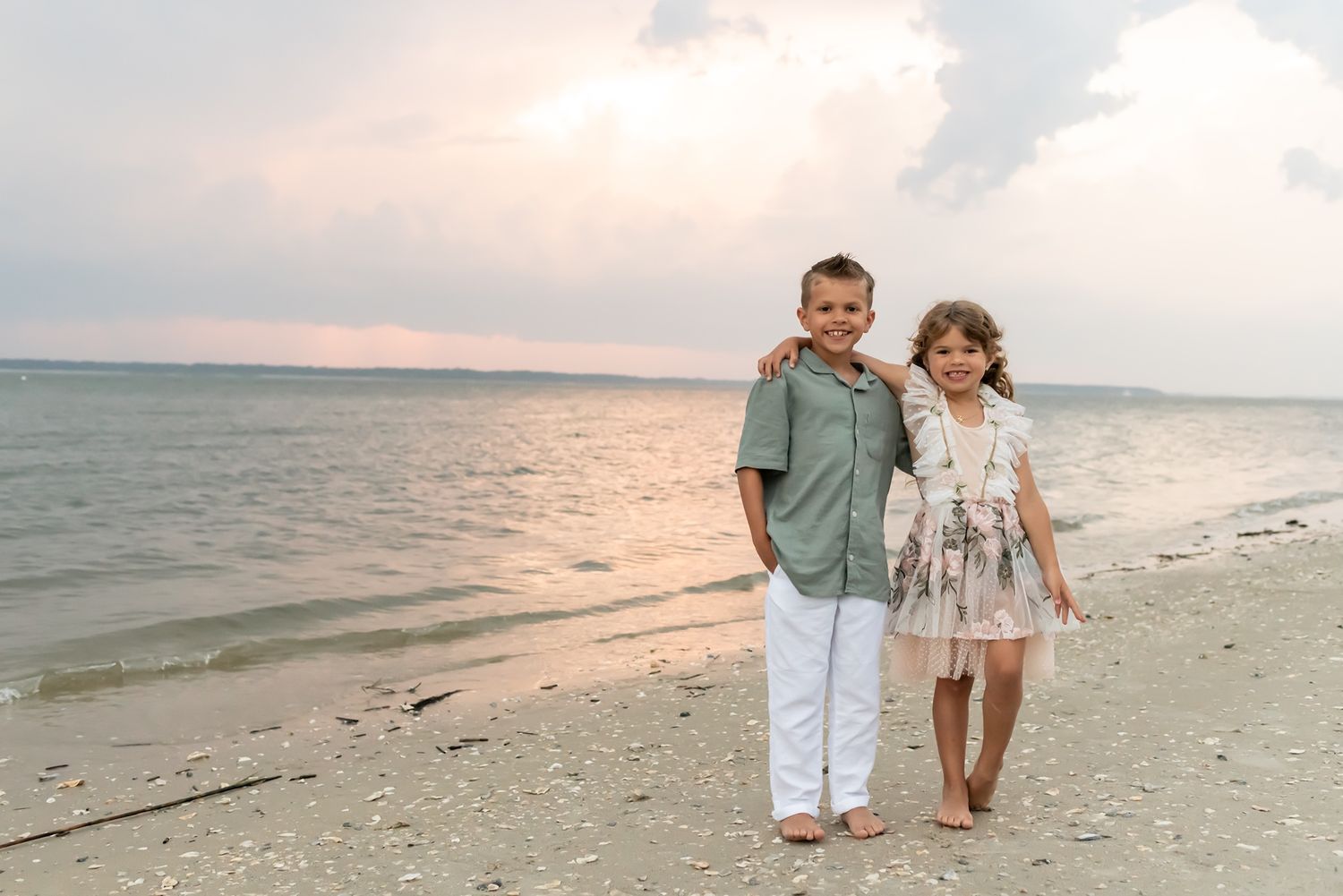 Two children stand together on a peaceful beach at sunset with calm ocean waters and cloudy sky in the background.