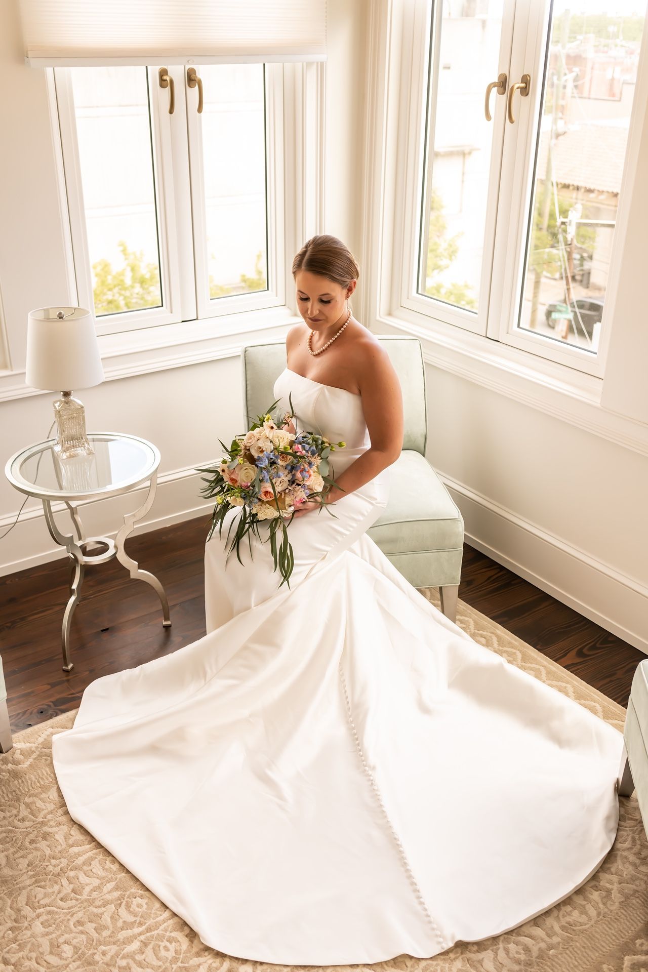 A stunning bride in a classic white ballgown poses gracefully by sunlit windows in an elegant room.