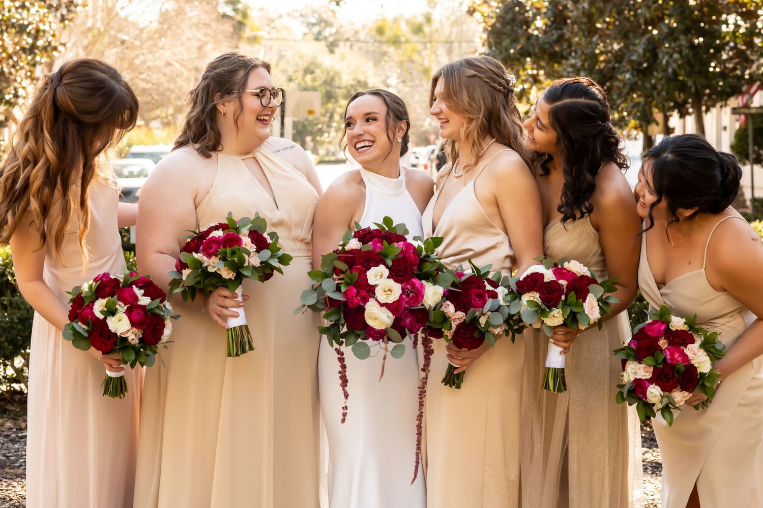 Joyful bridal party in blush pink bridesmaid dresses holding deep red and white floral bouquets outdoors.