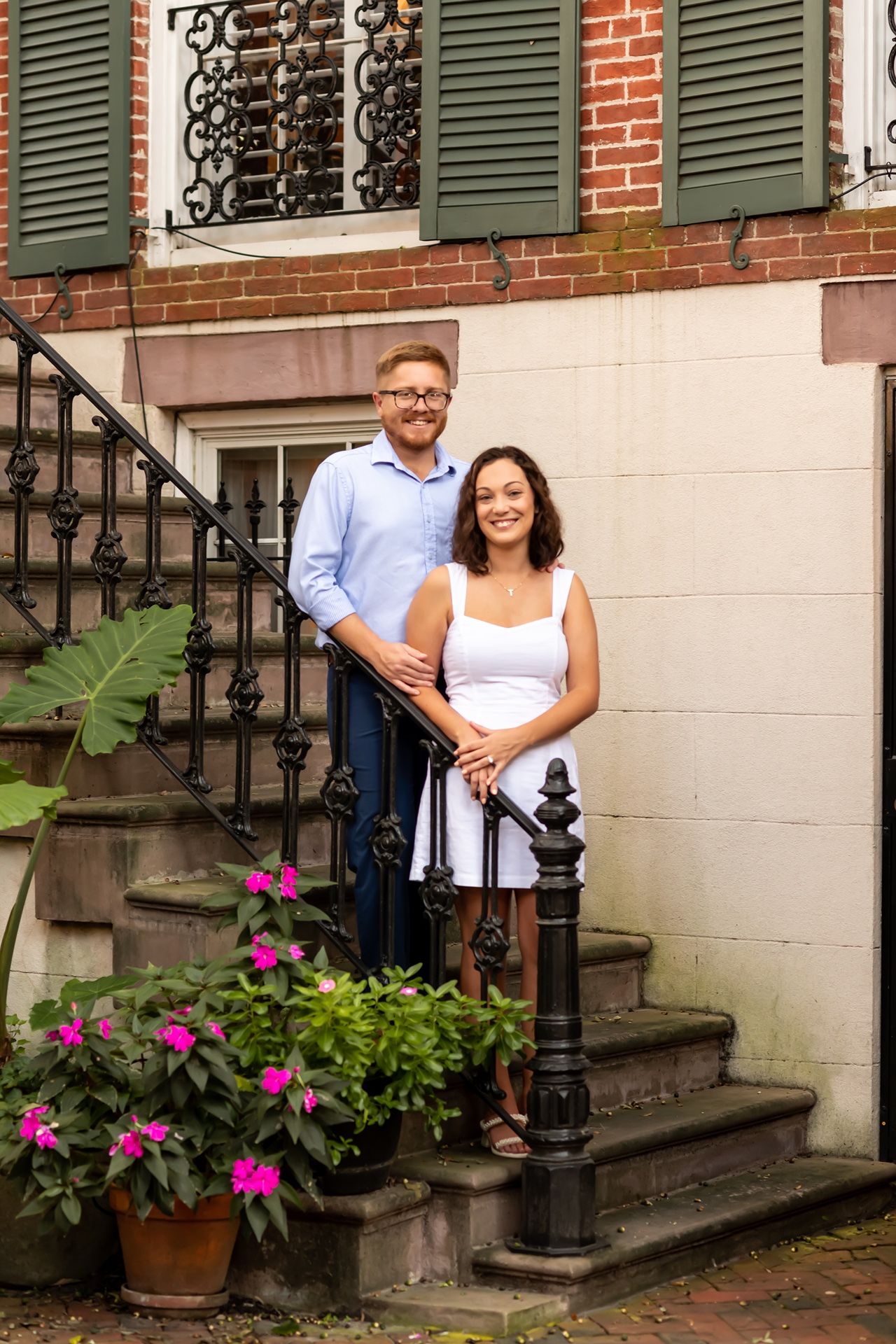 A couple stands together on brick townhouse steps decorated with bright pink flowers and green shutters.