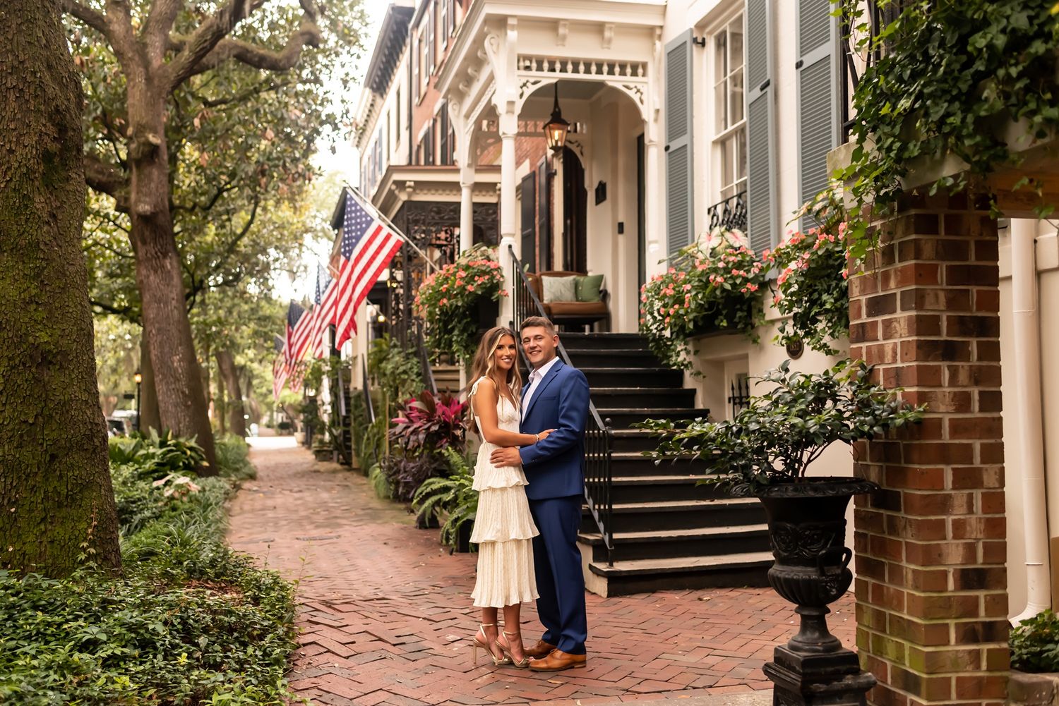 A couple stands on a brick sidewalk in front of a historic house with American flag and ornate Victorian porch details.