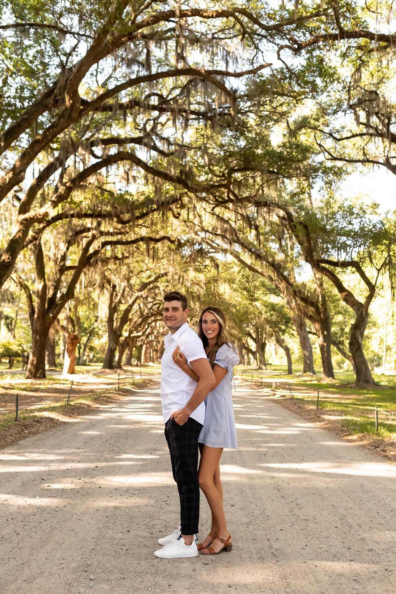 A couple embraces on a sunlit path beneath Spanish moss-draped oak trees in a romantic Southern setting.