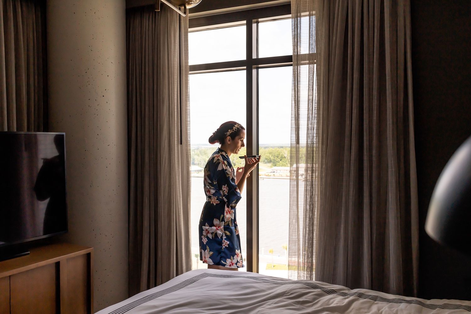 A silhouetted figure in a floral dress stands by a large window in a hotel room with flowing curtains.