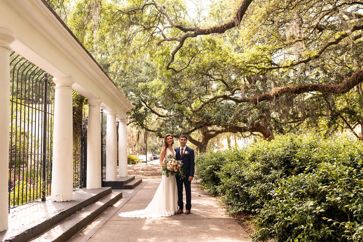 Two people stand under a grand oak tree with Spanish moss on a white-columned veranda walkway.