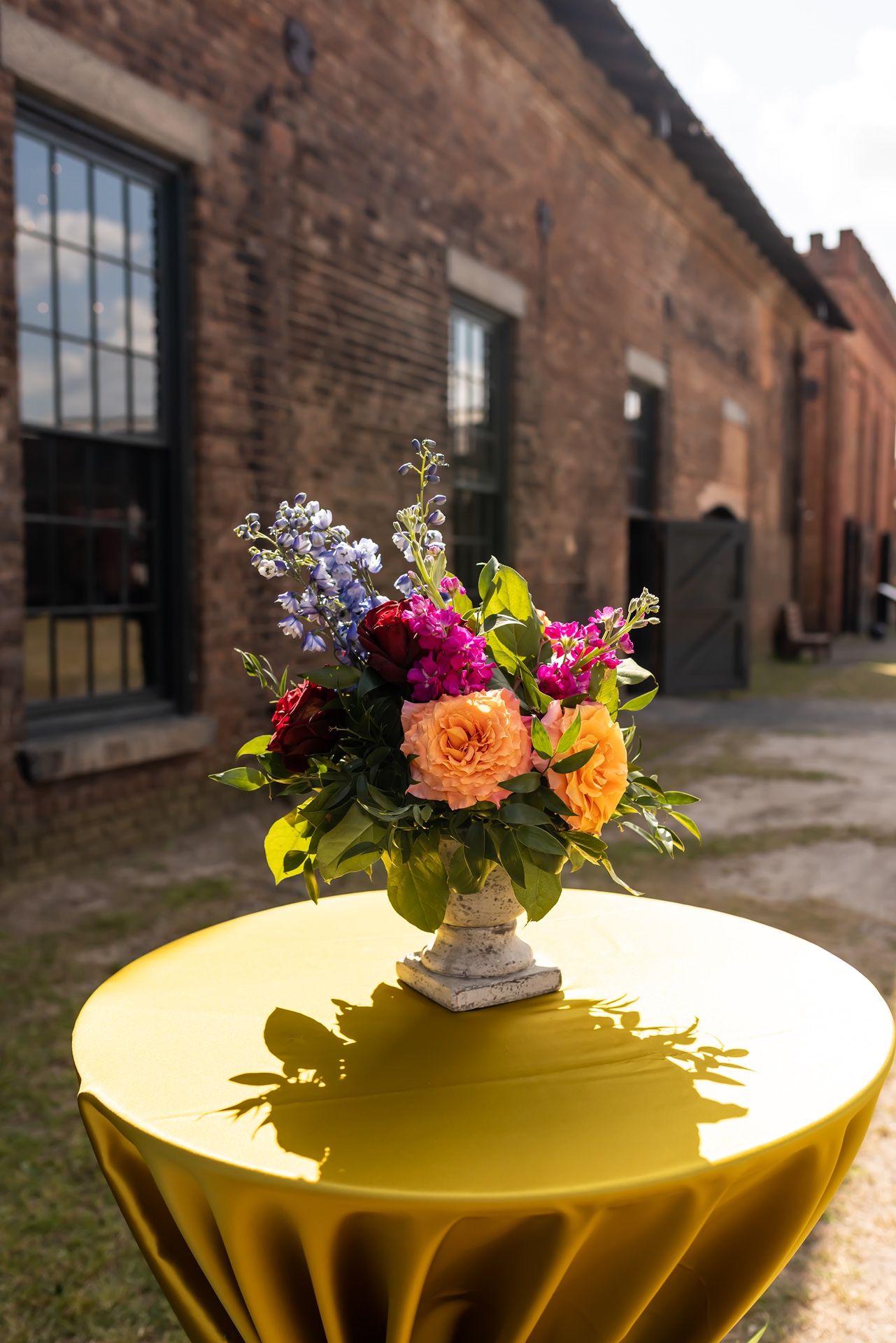 Colorful floral arrangement with roses and wildflowers sits on yellow table against rustic brick building exterior.