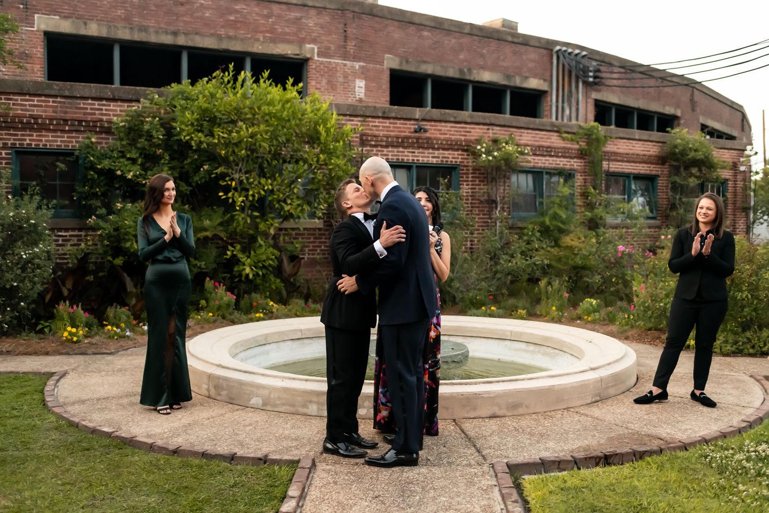 A wedding ceremony takes place by a circular fountain with brick buildings and garden flowers in the background.