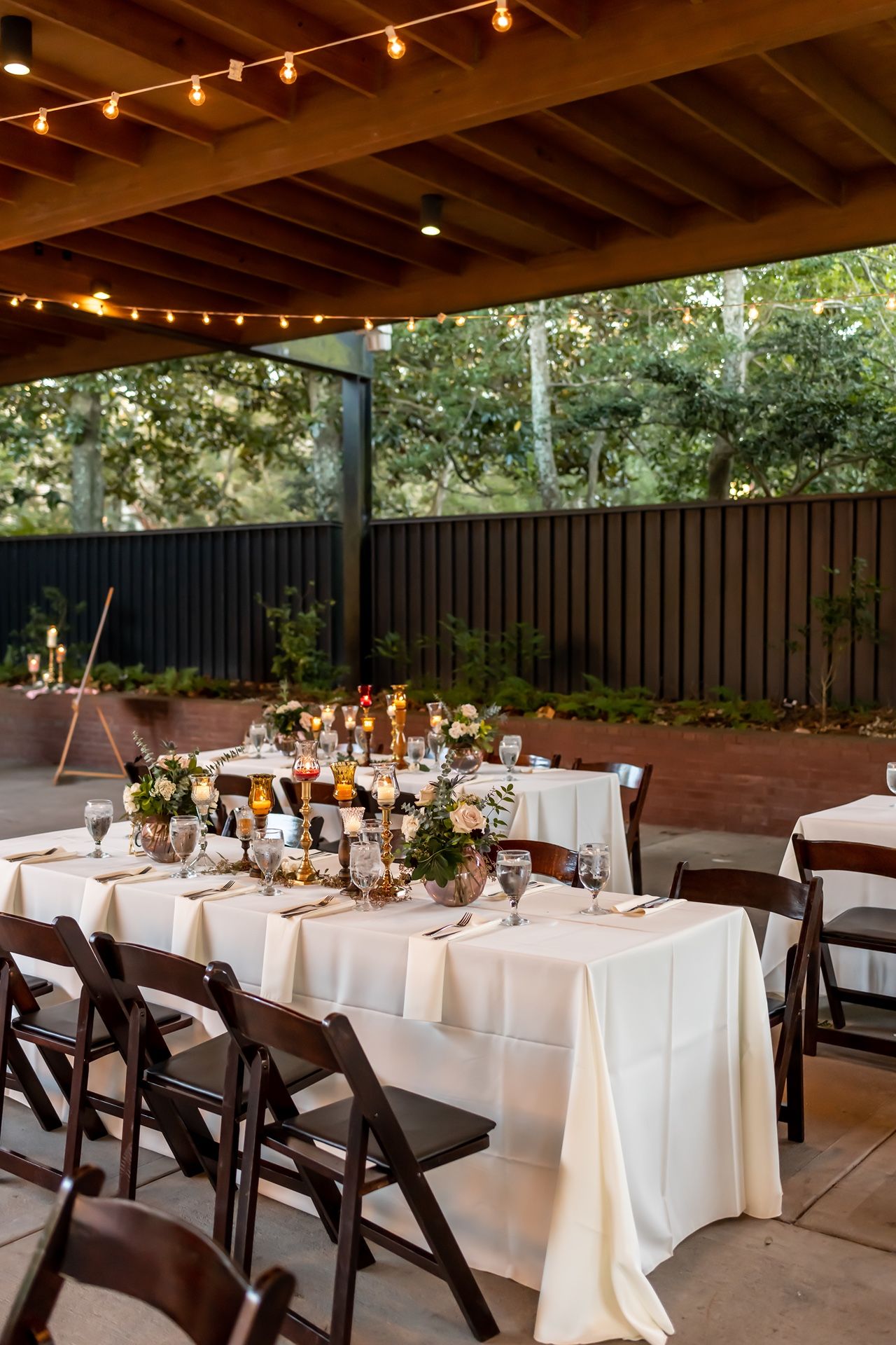 Wedding reception dining table set with white linens and string lights on covered outdoor patio with greenery backdrop.