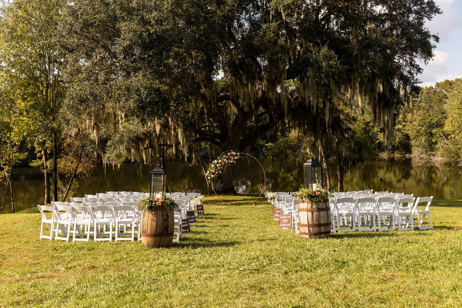 Elegant outdoor wedding ceremony setup with white chairs and floral arrangements under Spanish moss draped trees.