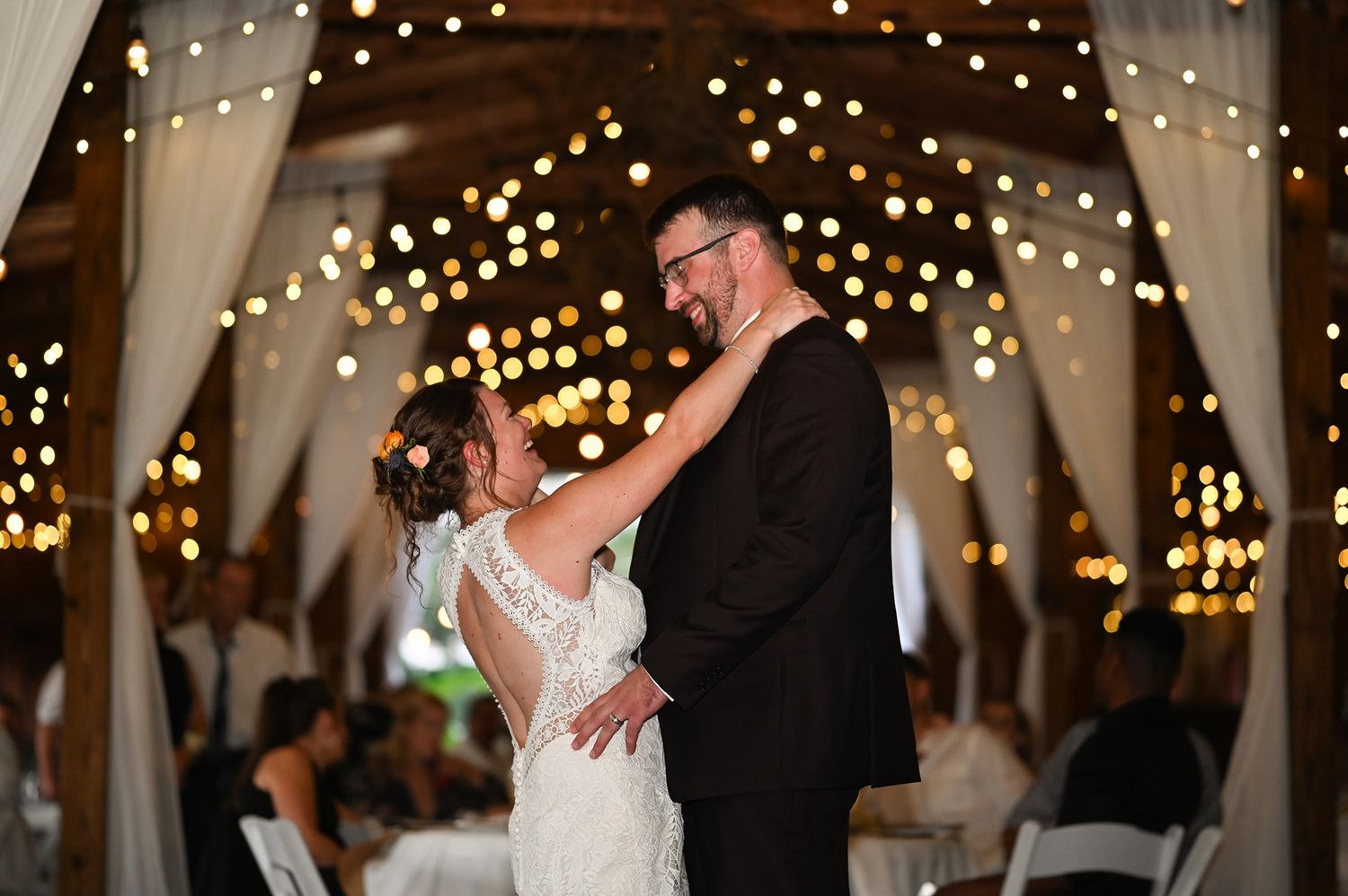A couple shares their first dance at a wedding reception beneath twinkling string lights and draped fabric decor.