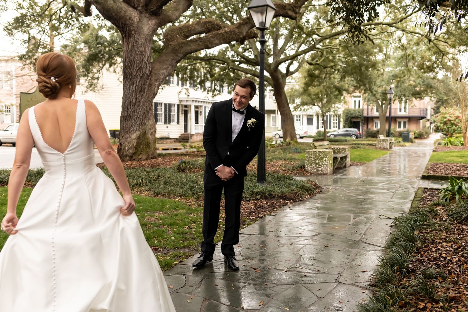 A couple shares their first look moment on a rainy garden path beneath moss-draped oak trees in a Southern garden setting.