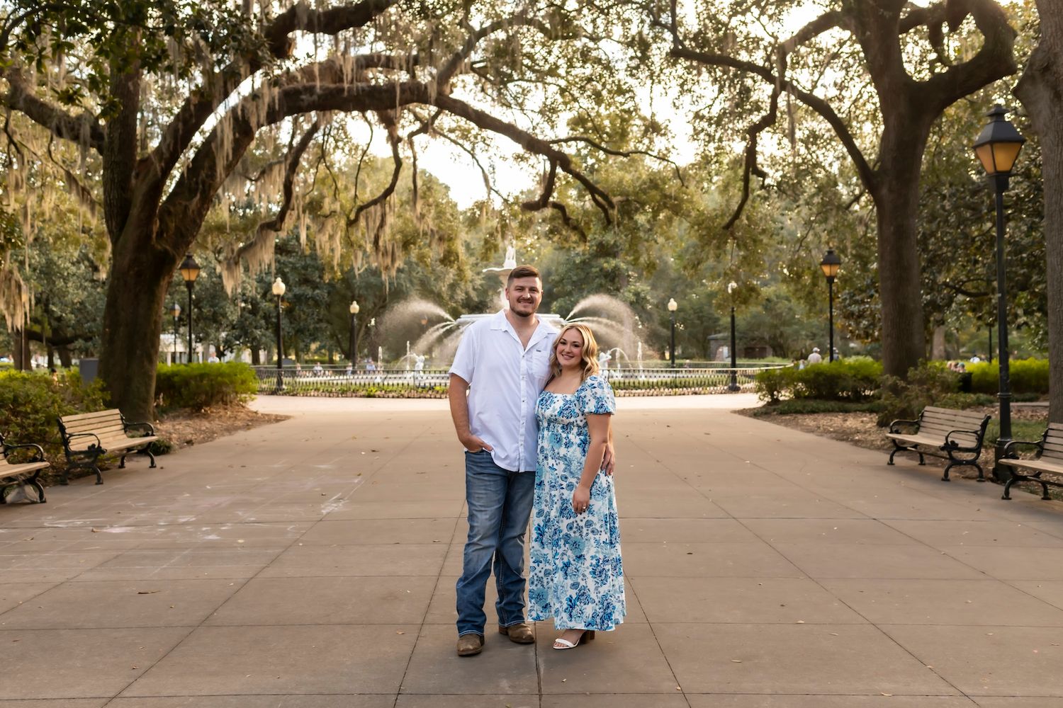 A couple stands together on a paved walkway under oak trees draped with Spanish moss in a scenic park setting.