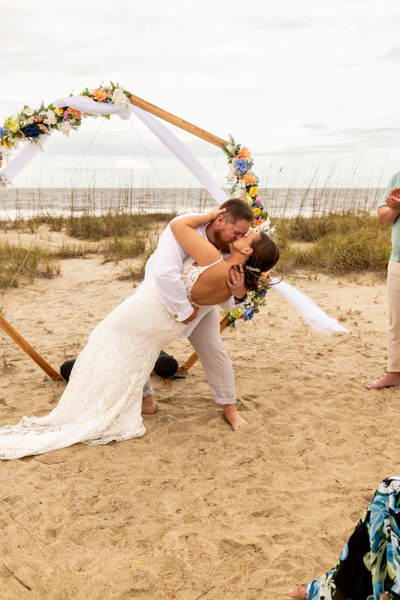 A romantic beach wedding ceremony with a wooden arch decorated with flowers and flowing fabric on a sandy shore.